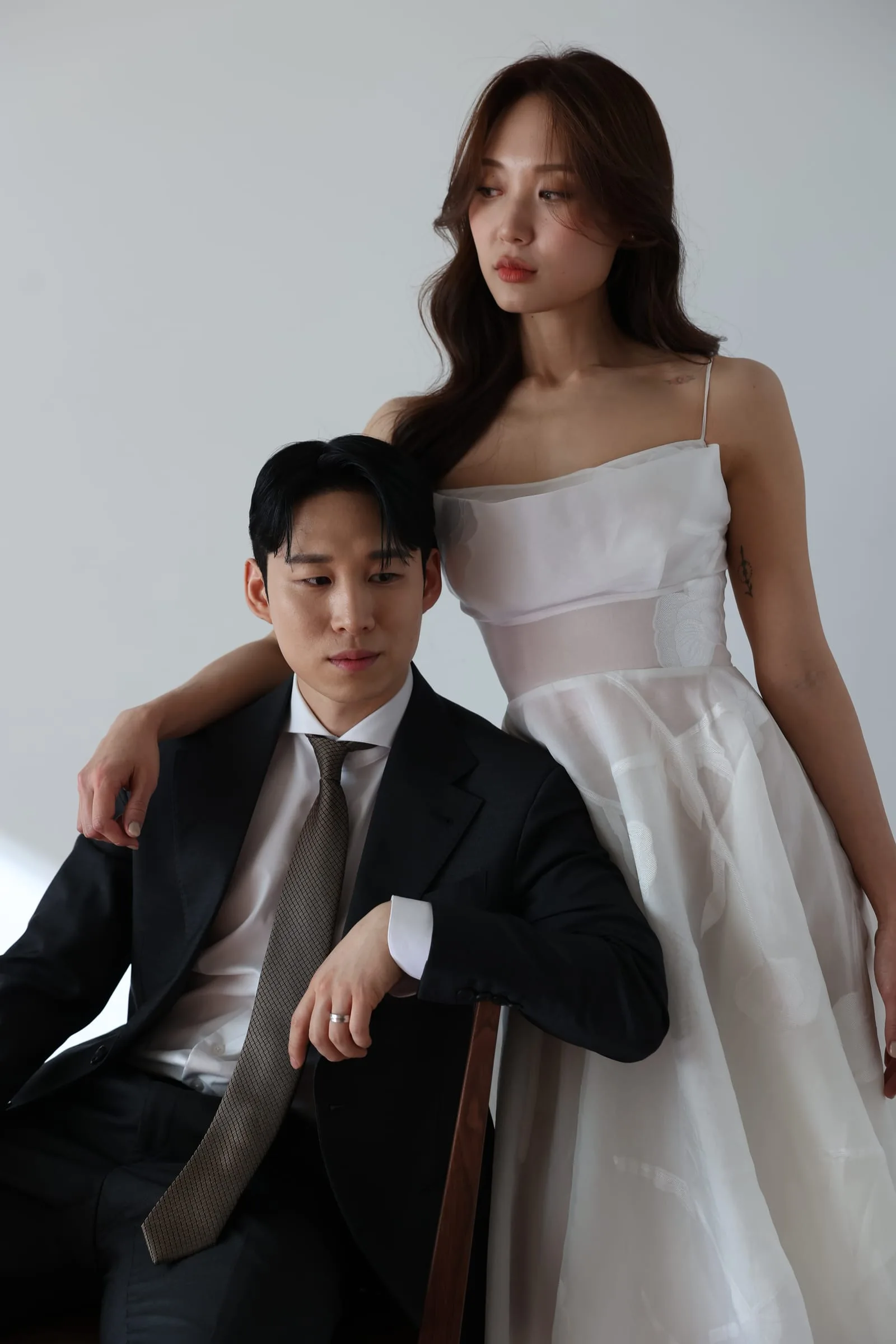 컬러 스튜디오 — 의자에 앉은 예복 신랑의 어깨에 신부가 팔을 얹고 선 결혼 화보