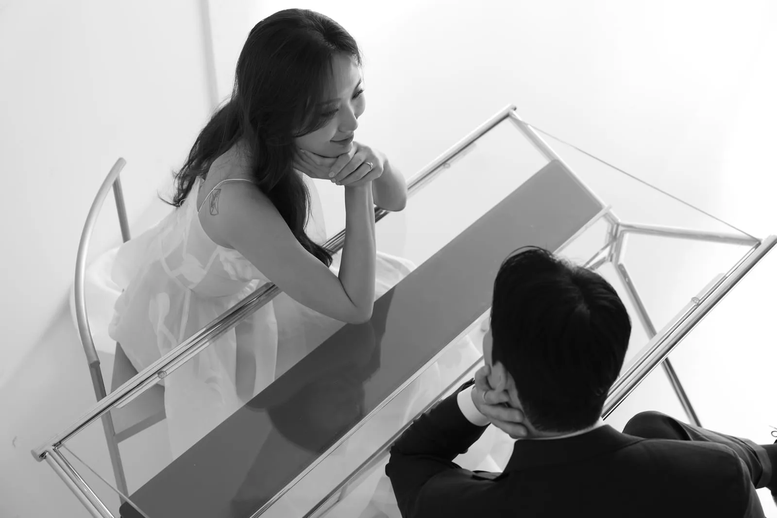 흑백 부감 — 유리 테이블을 사이에 두고 마주 앉은 신랑·신부의 결혼 스튜디오 사진