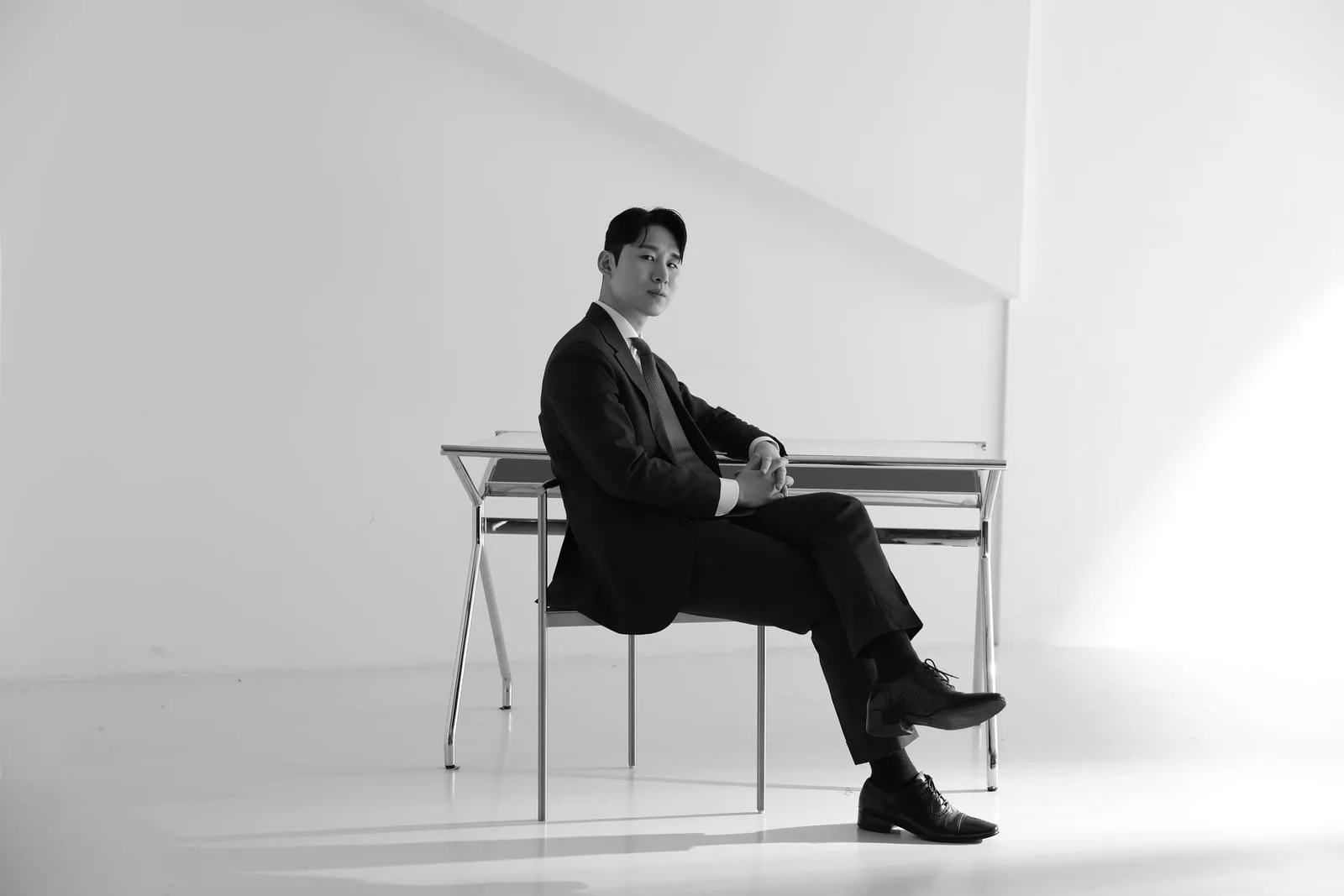 흑백 전신 — 금속 벤치에 기댄 채 카메라를 응시하는 신랑, 맞춤 다크 수트와 드레스 슈즈