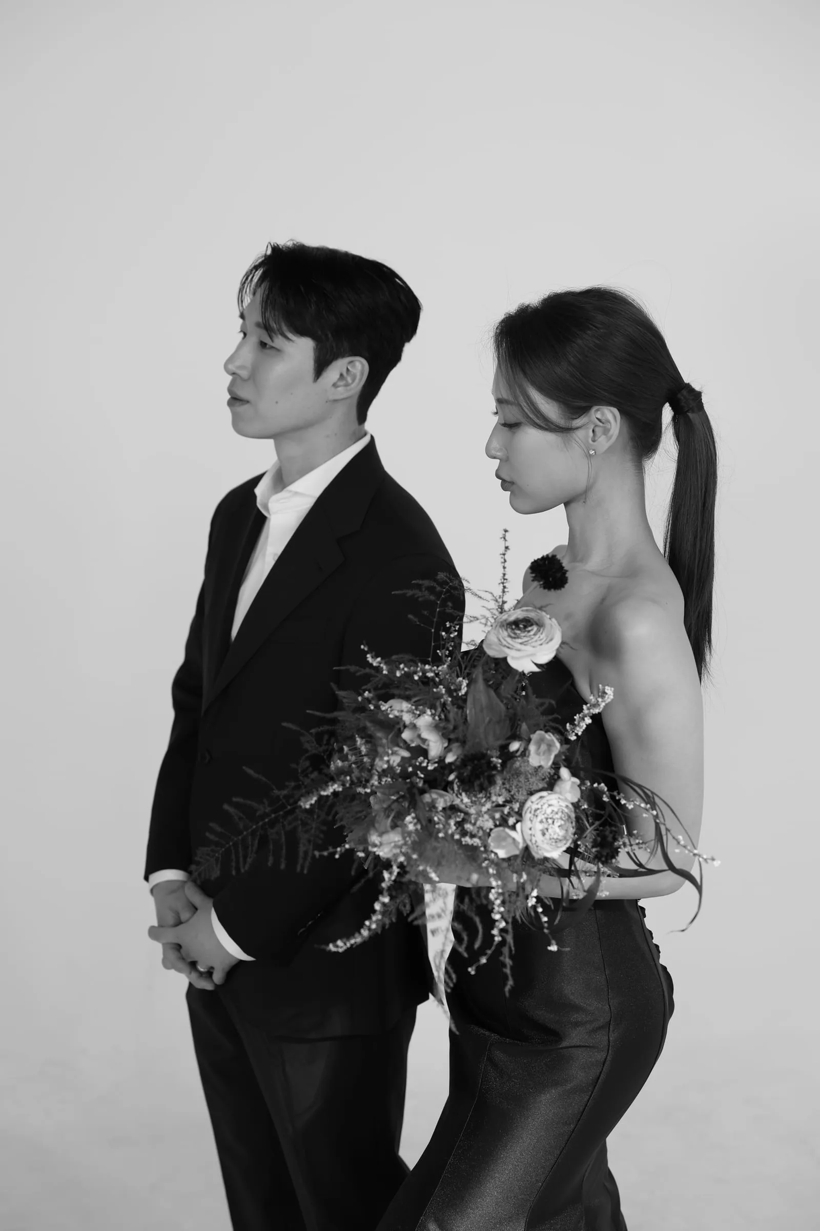흑백 스튜디오 — 다크 수트 신랑과 꽃다발 든 신부가 나란히 선 결혼 사진 옆모습