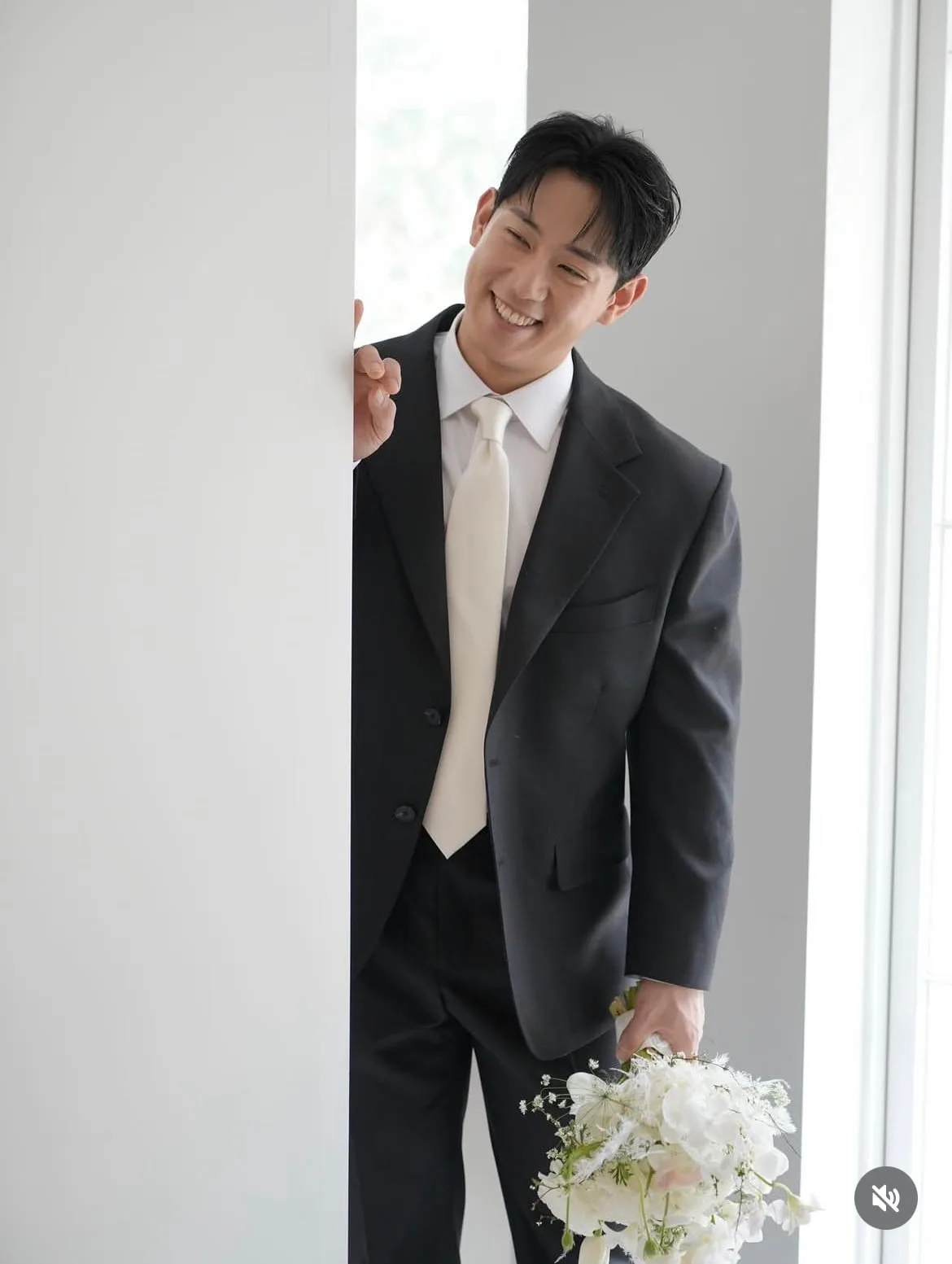 컬러 — 차콜 수트에 아이보리 타이를 맨 신랑이 흰 꽃다발을 들고 활짝 웃으며 벽에 기댄 결혼 스냅