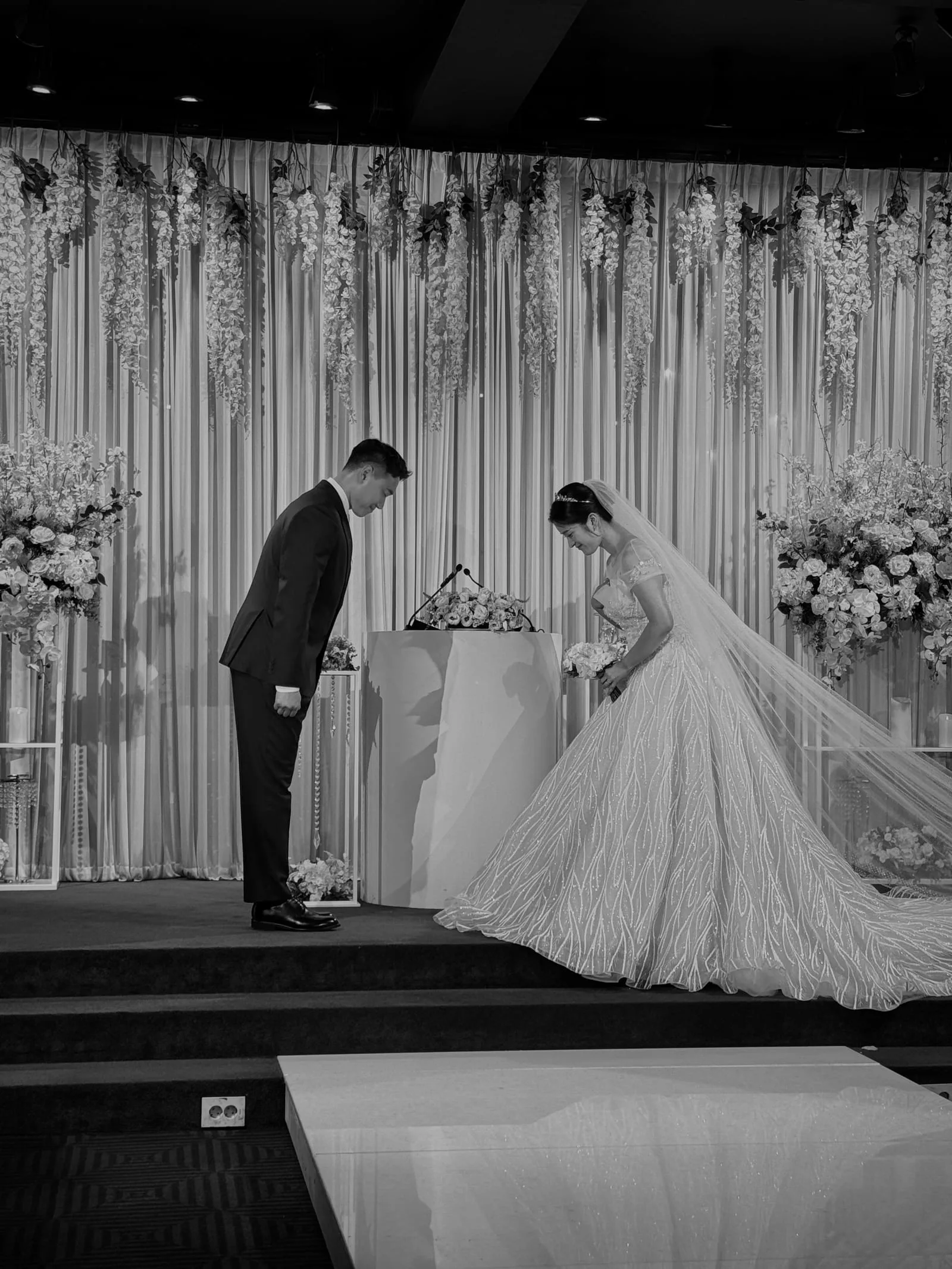 흑백 예식장 — 다크 수트 신랑과 볼가운 신부가 예식 제단 앞에서 인사를 나누는 순간