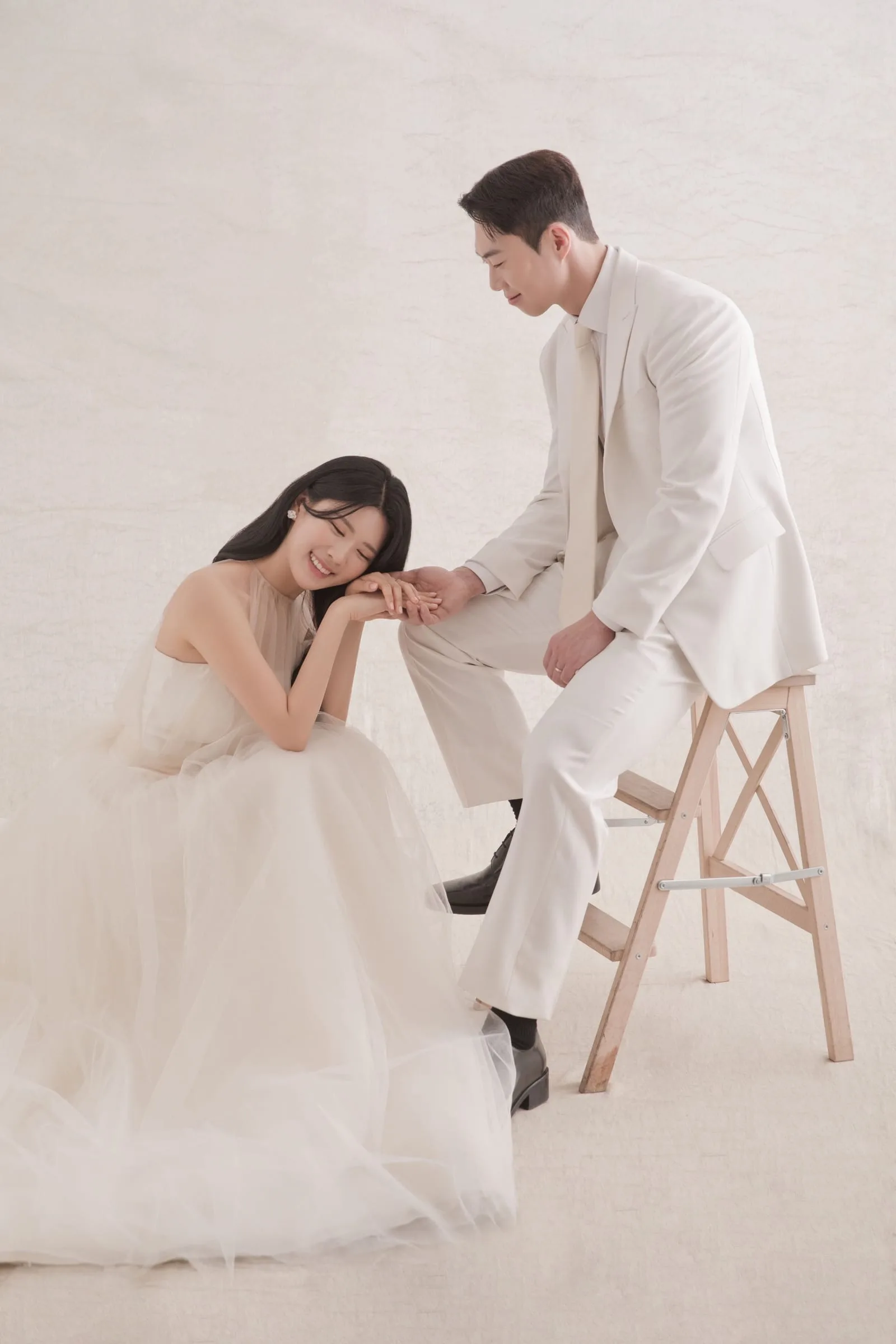 컬러 밝은 스튜디오 — 아이보리 수트 신랑과 흰 드레스 신부가 손을 맞잡고 마주 앉은 웨딩 화보