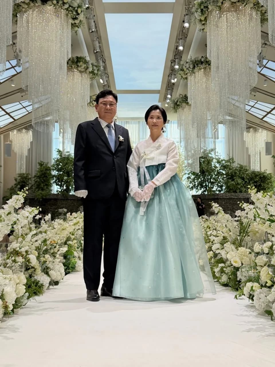 컬러 — 꽃 장식 예식장 아일에 선 혼주 부부, 아버지 다크 혼주복, 어머니 민트 한복