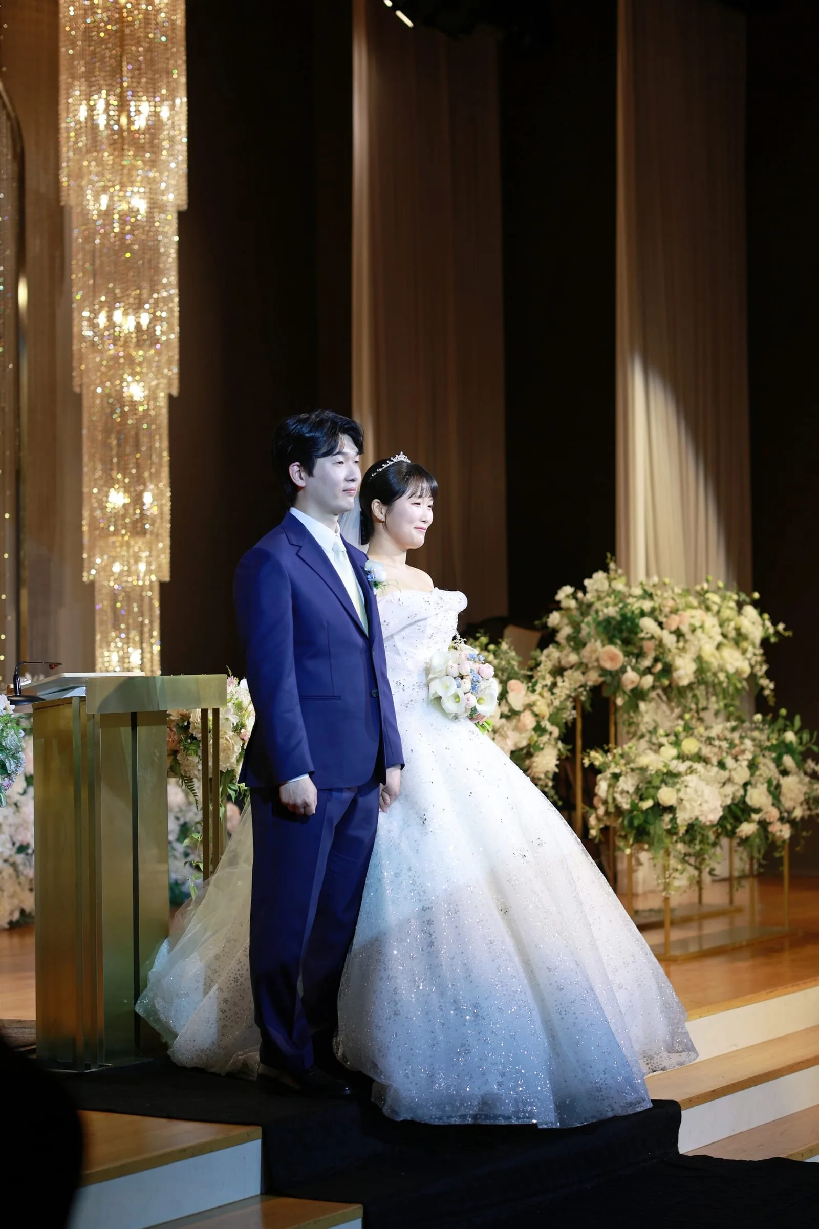 컬러 예식장 — 블루 예복 수트 신랑과 반짝이 볼가운 신부가 황금빛 샹들리에 아래 예식 스테이지에 나란히 선 모습
