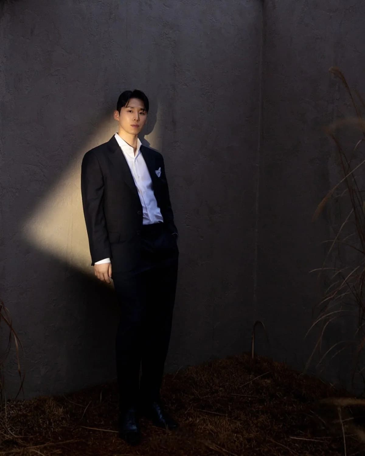 컬러 무드 — 콘크리트 벽에 기댄 노타이 다크 수트 신랑, 드라마틱한 빛 줄기와 억새 배경의 예복 화보
